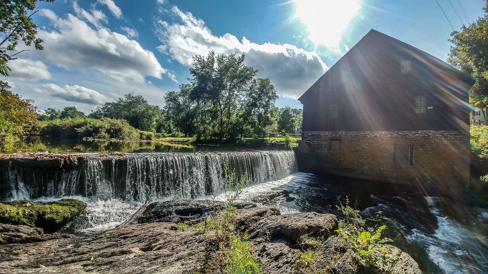 A waterfall is surrounded by trees and a building with the sun shining through the clouds.