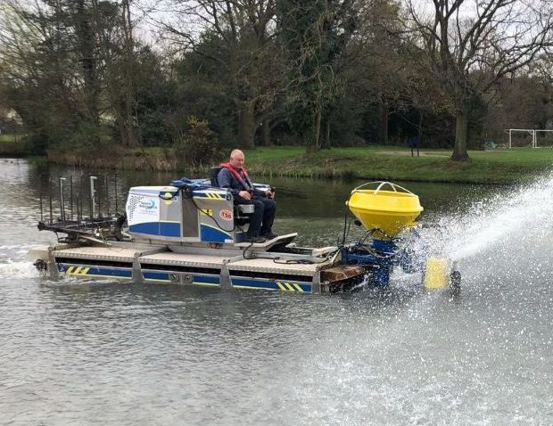 Man operating a blue and yellow aquatic weed harvester on a lake, spraying water.
