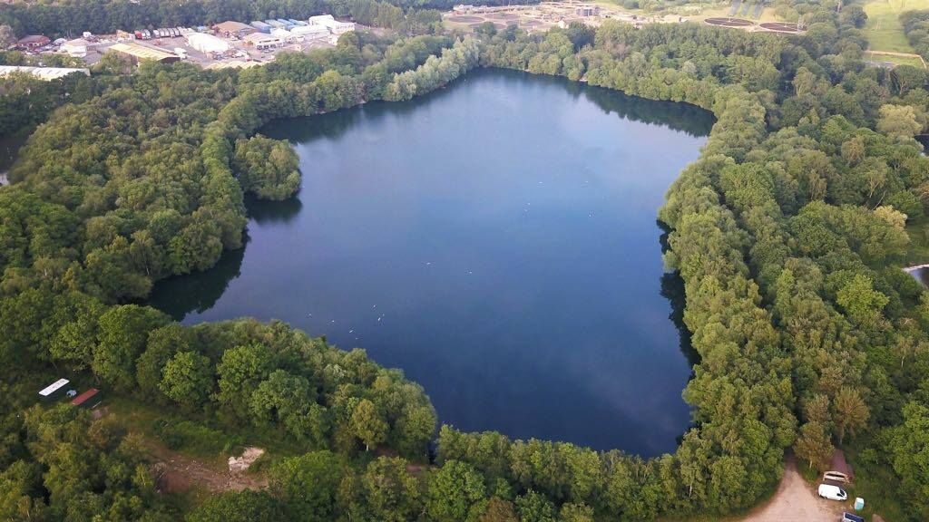 Aerial view of a dark lake surrounded by lush green trees, with a sandy area on one side.
