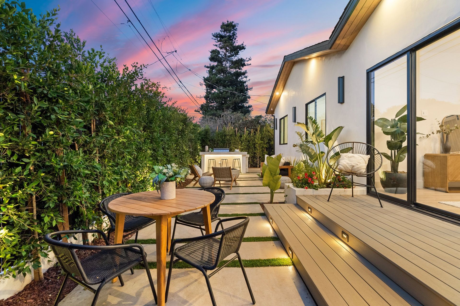 Patio moderne dans le jardin avec table ronde, chaises, terrasse en bois, plantes en pot et ciel au coucher du soleil