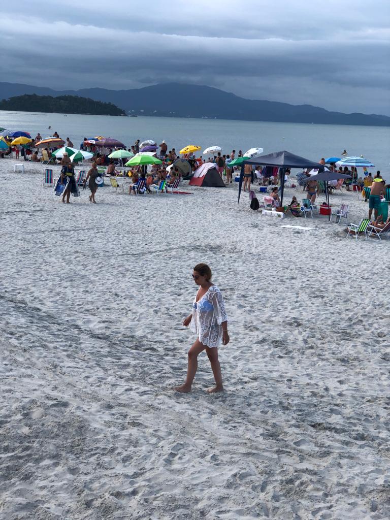 Uma mulher caminha em uma praia cercada por pessoas e guarda-sóis.