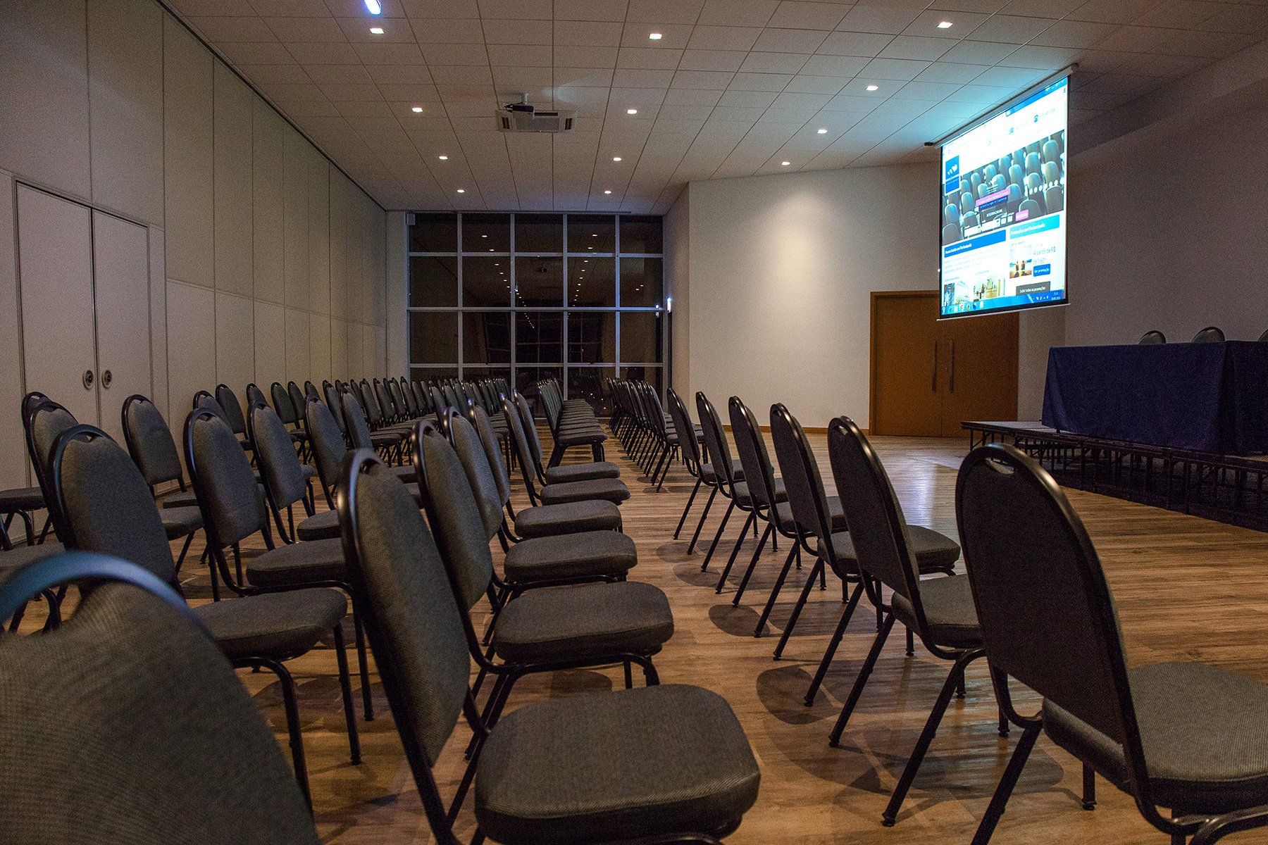 Fileiras de cadeiras estão alinhadas em uma sala de conferências com uma grande tela pendurada no teto.