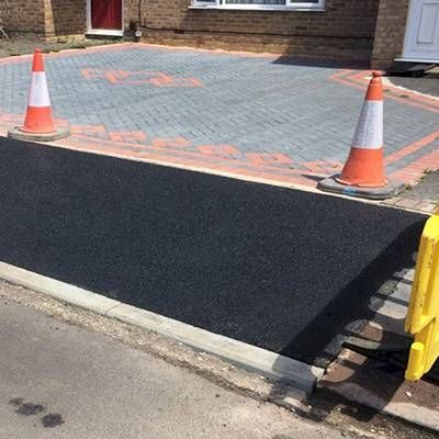 A black tarmac dropped kerb to a driveway with orange and white traffic cones in front of a brick house.