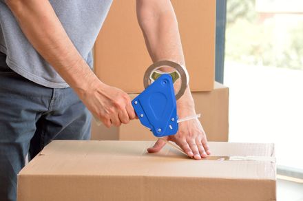 Hands using a blue tape dispenser to seal a cardboard box on a packing table.