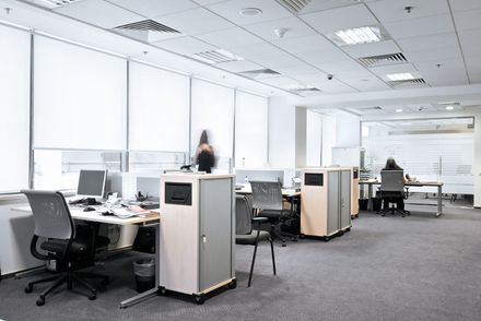 Bright office with desks, computer monitors, chairs, and two people working near windows