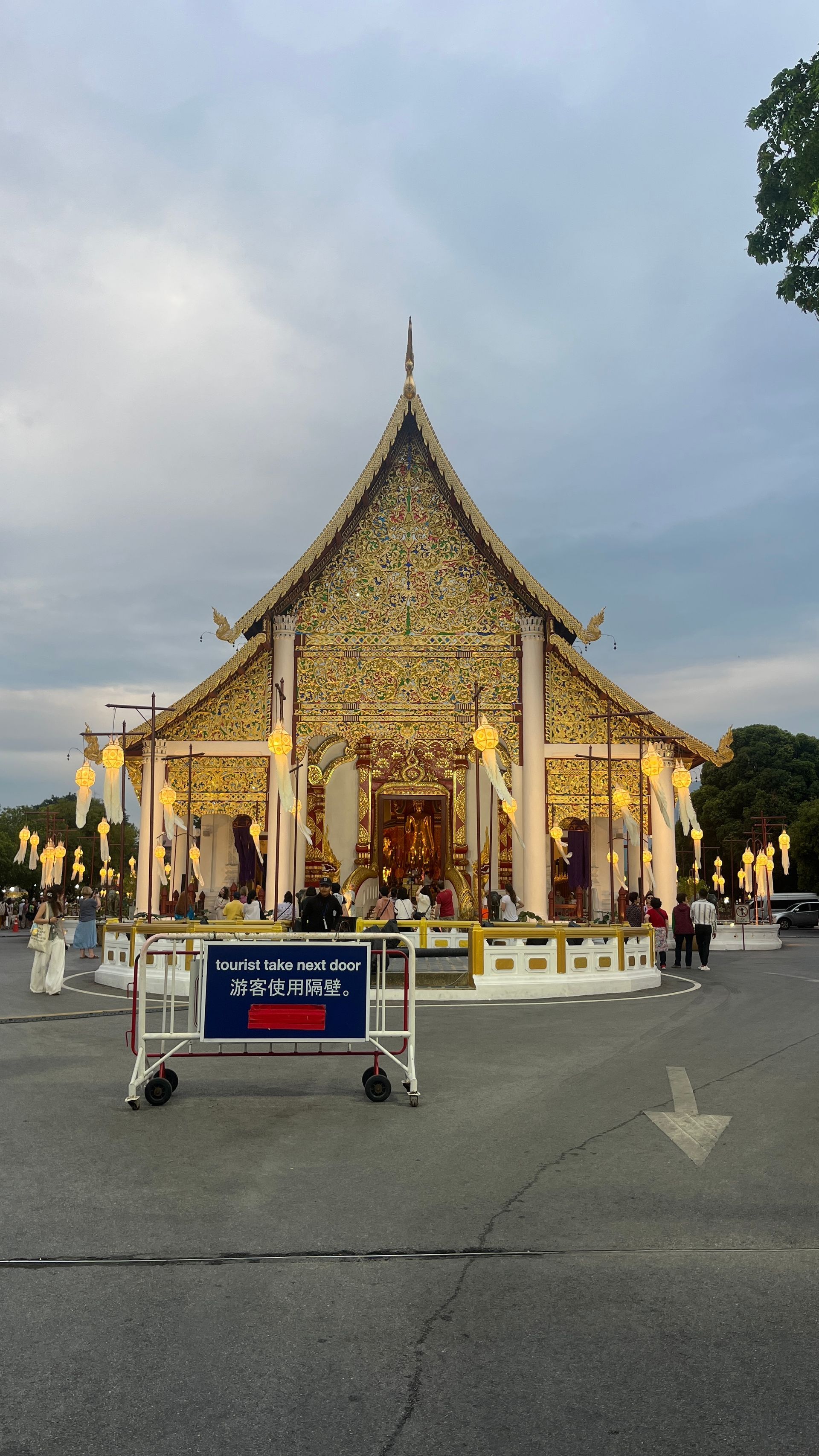 A decorative gold-gilded Thai temple building under a cloudy sky, with a white portable crowd barrier in the foreground.