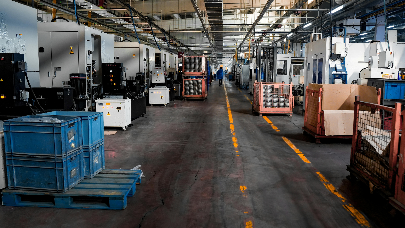 Factory floor with industrial machinery and a worker moving items in a metal container.