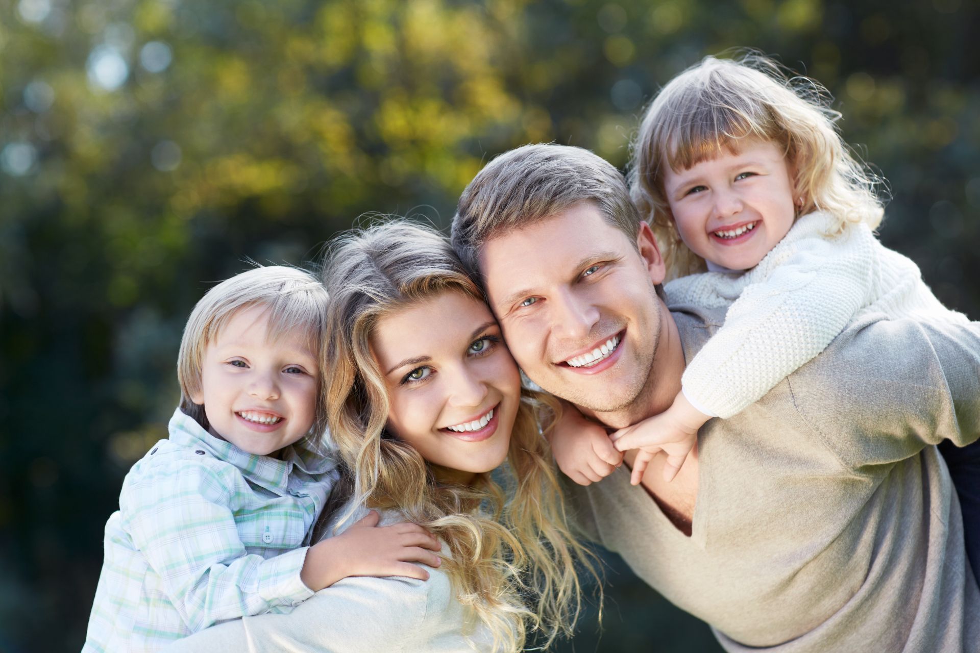 Family of four smiling, posing outdoors; father carrying daughter, mother holding son.