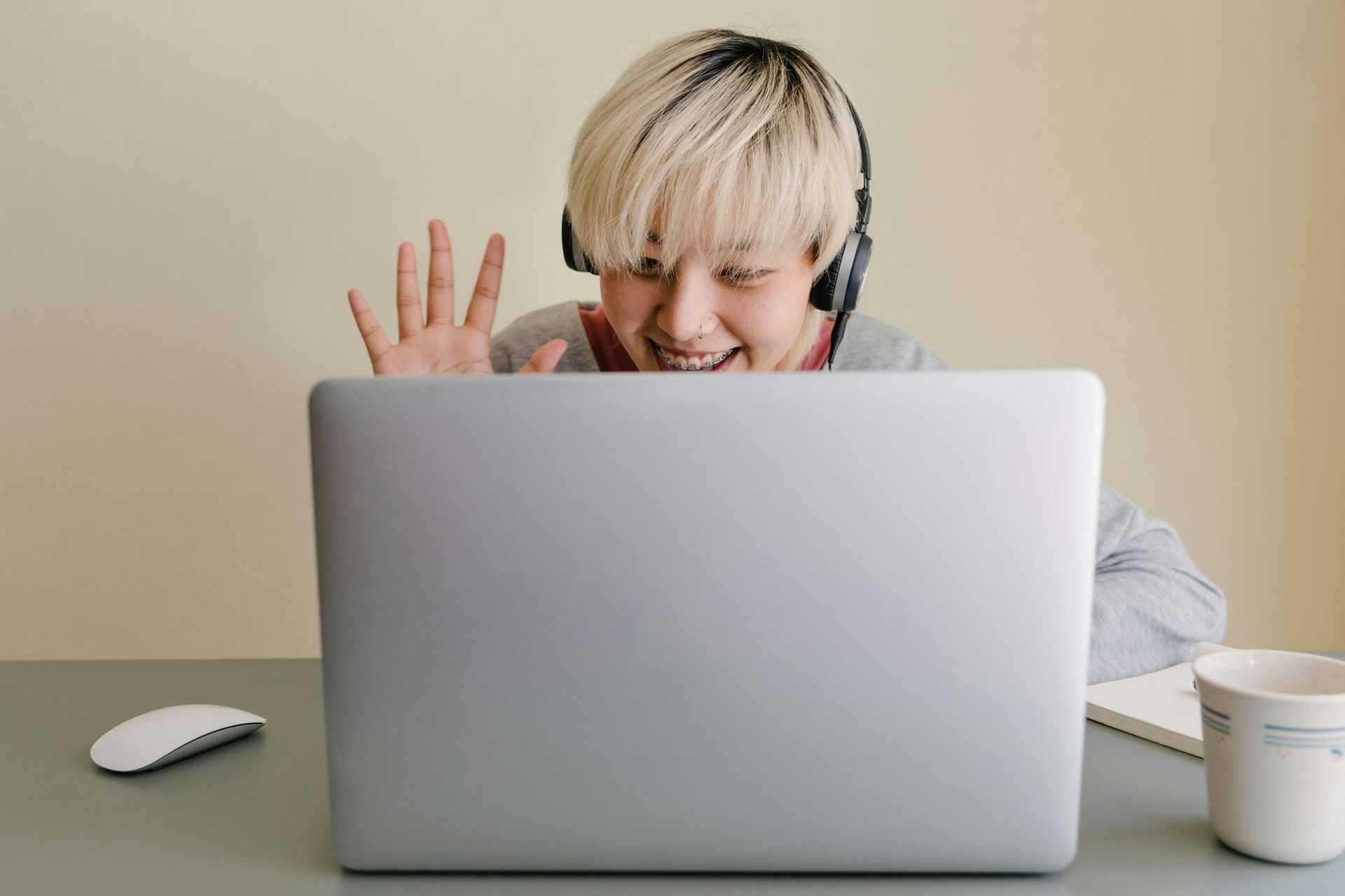 Person with headphones waves enthusiastically at laptop during video call. Light hair, neutral background.