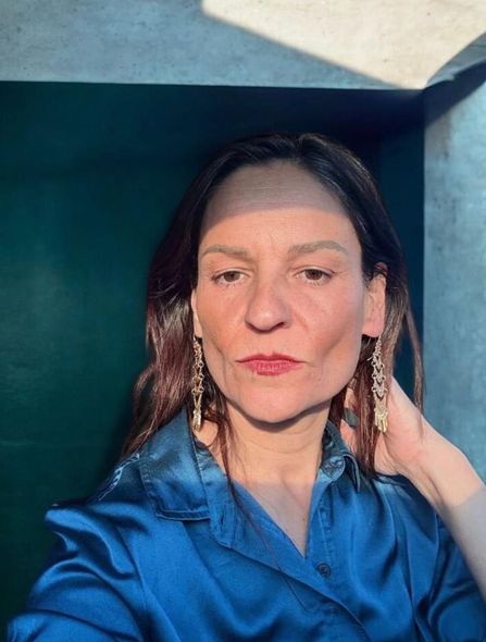 Woman in a blue satin shirt, wearing earrings, standing in sunlight by a green wall.