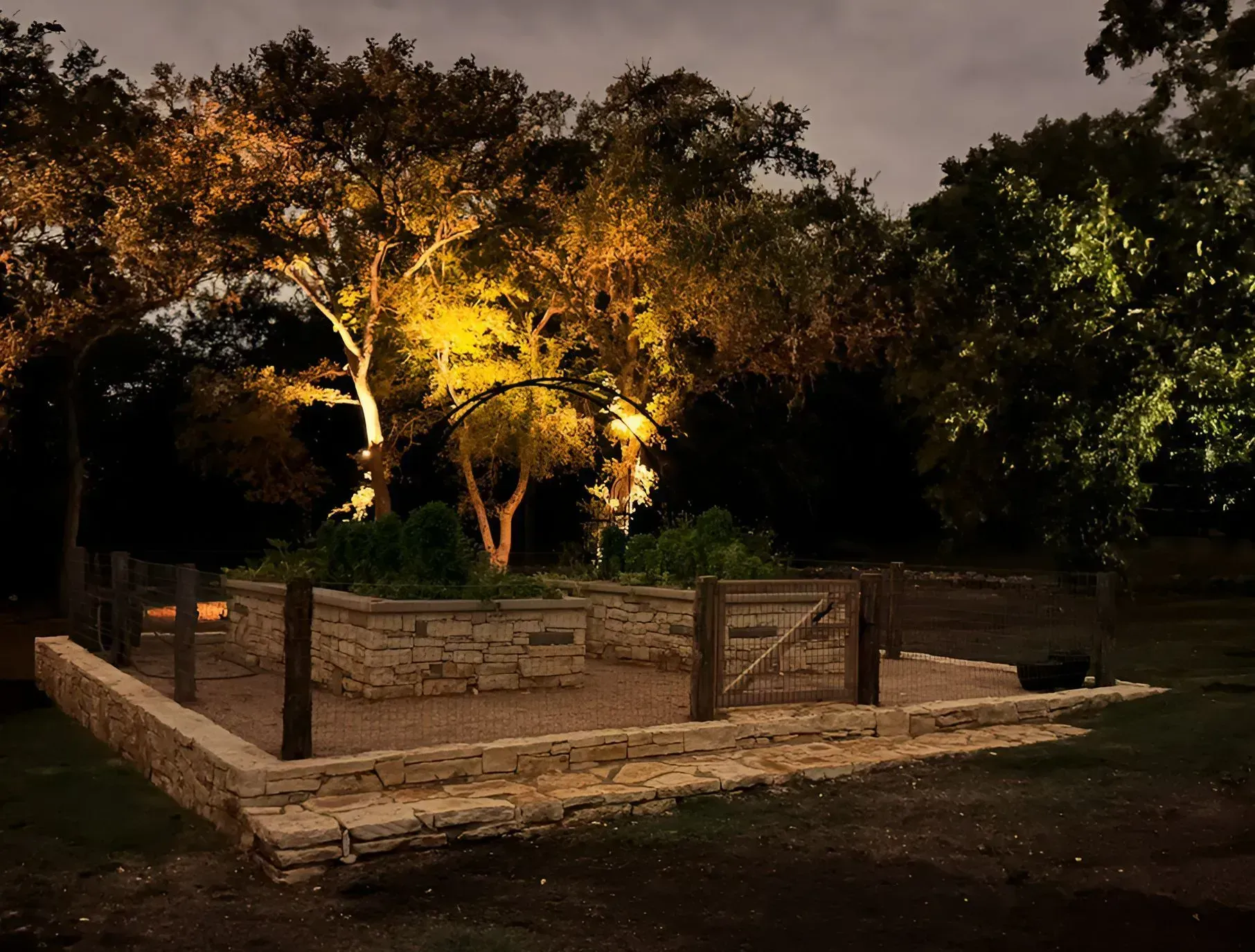 Nighttime view of a stone-walled garden bed with a wooden gate, illuminated by spotlights, trees in the background.