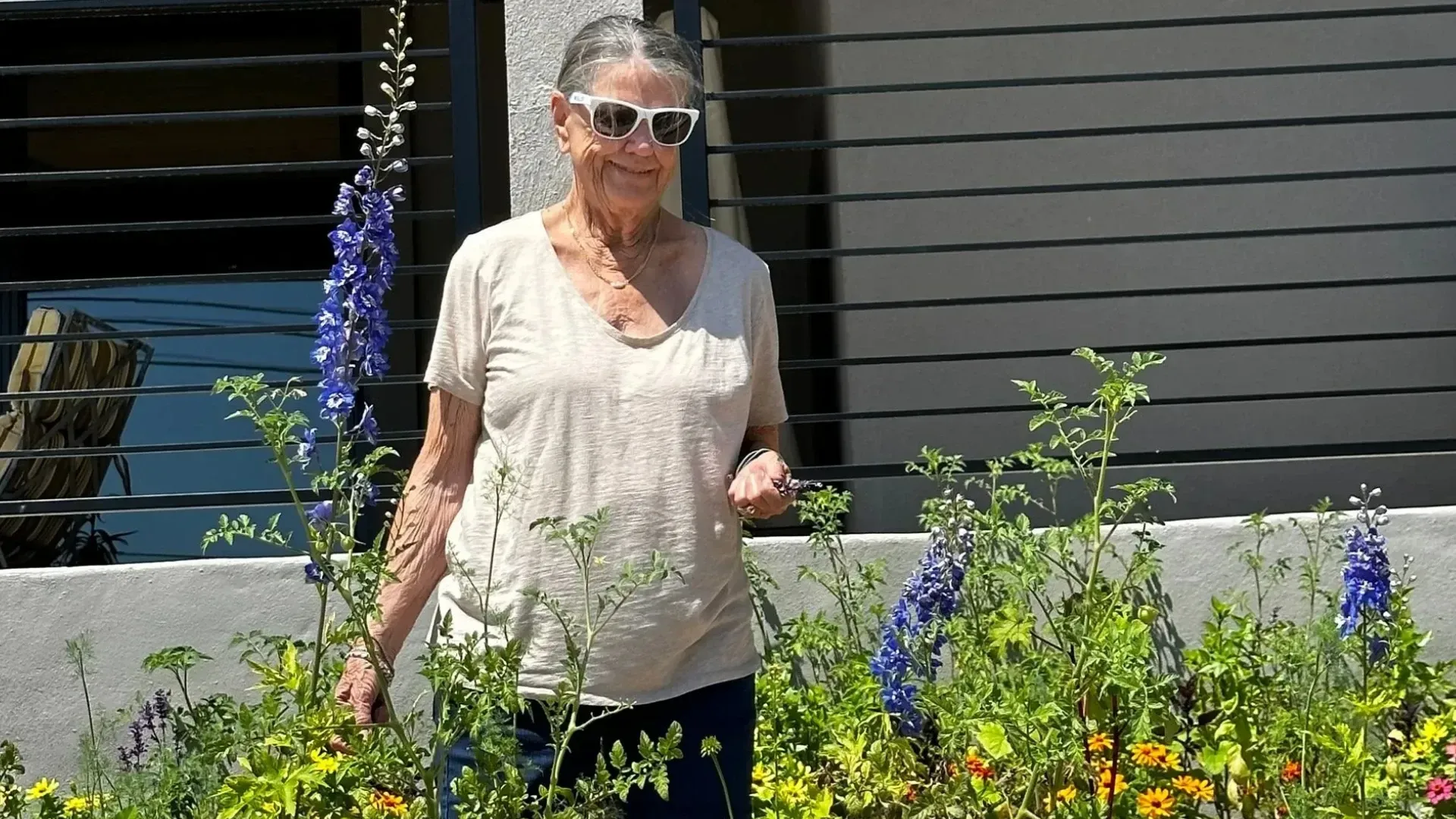 Woman in sunglasses standing in a flower bed, near a building.