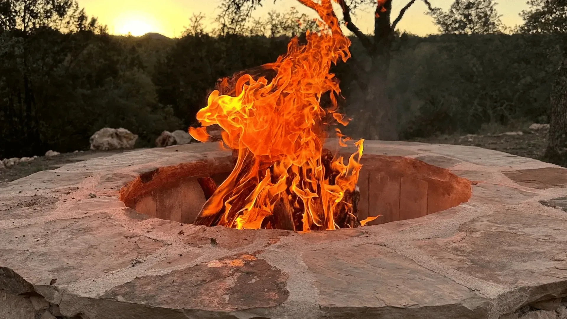Fire pit with bright orange flames, stone structure, sunset backdrop.