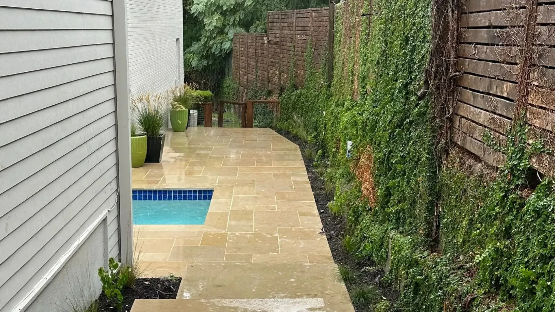 Stone pathway leads past a house and a pool, alongside a wooden fence covered in greenery.