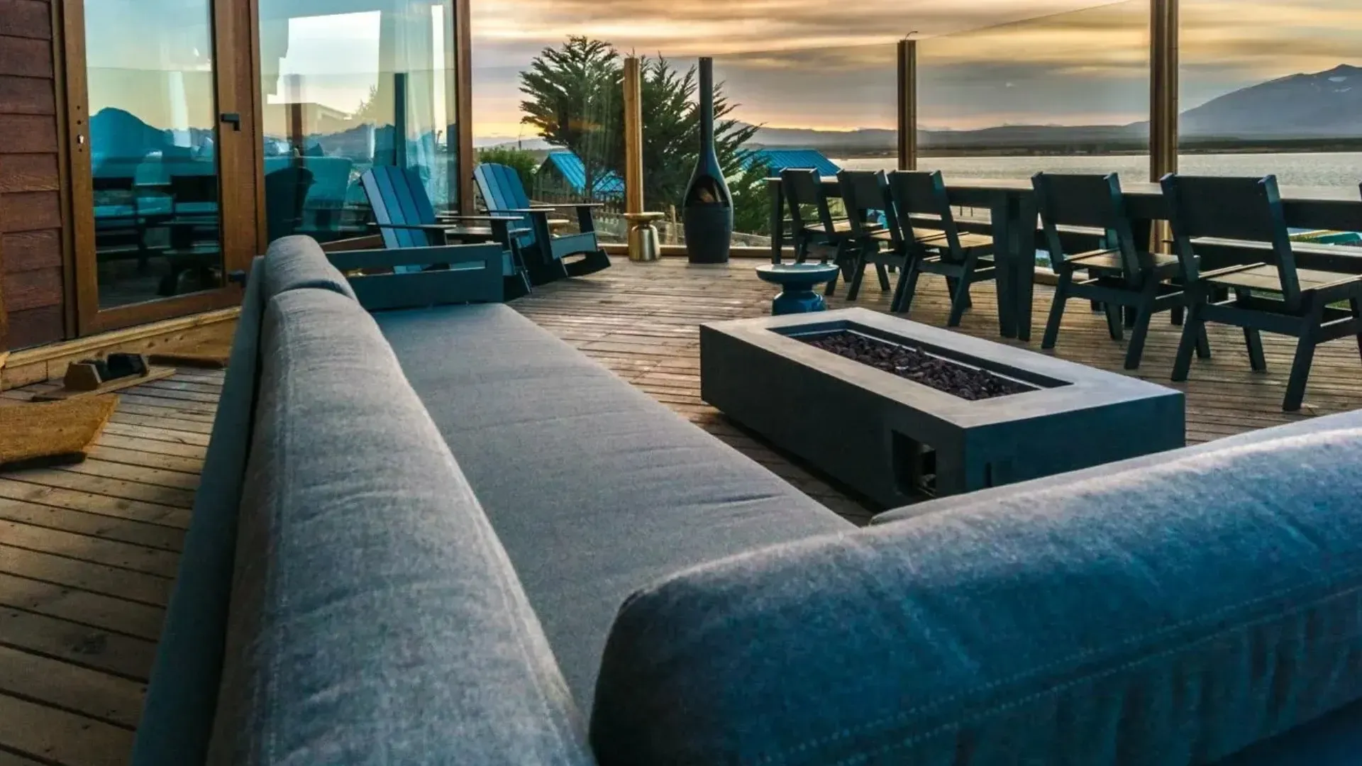 Deck of a modern home overlooking water at sunset. Outdoor sofa, dining table, and fire pit.