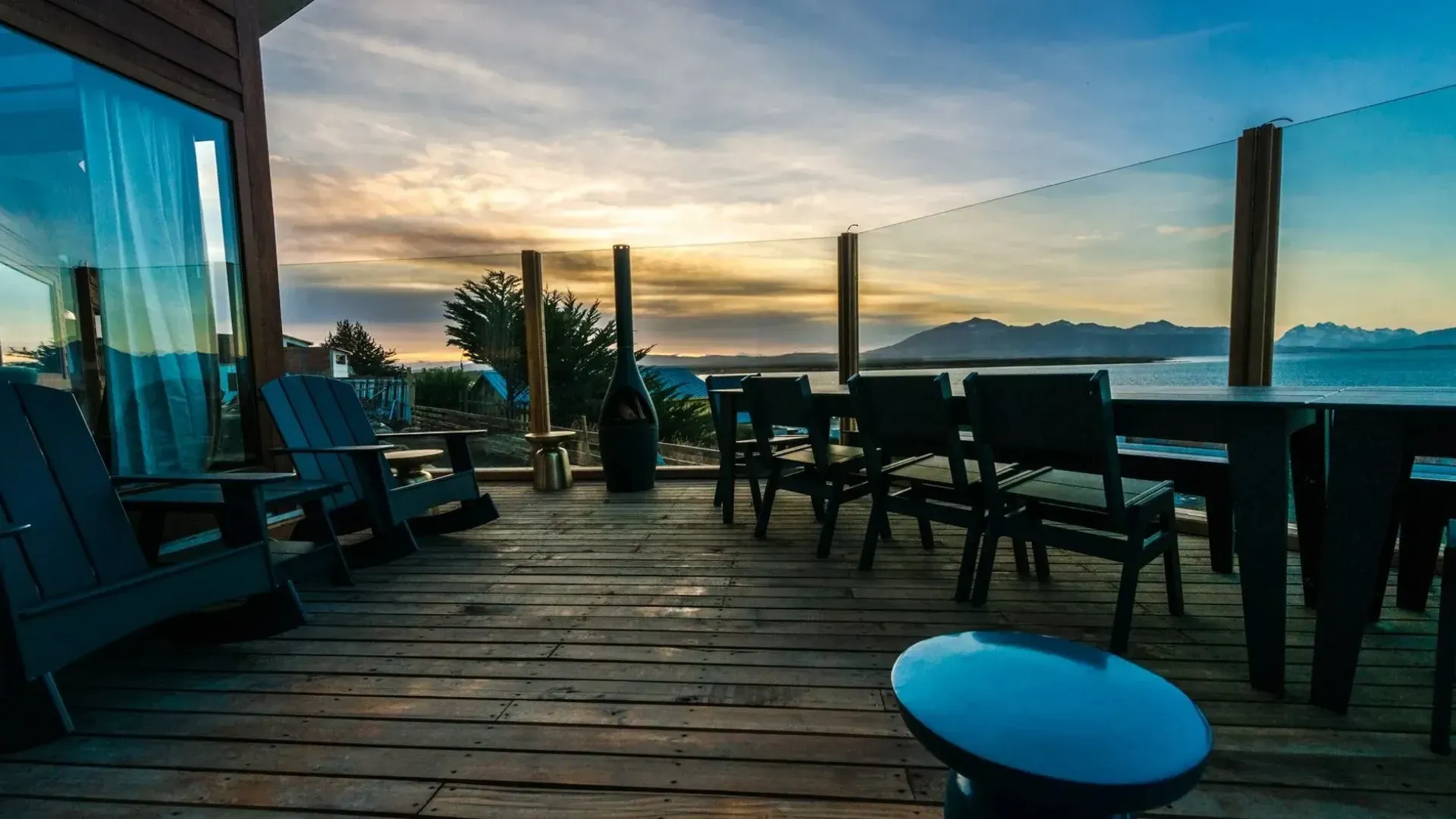 Wooden deck with outdoor furniture, glass railing, and water view at sunset.