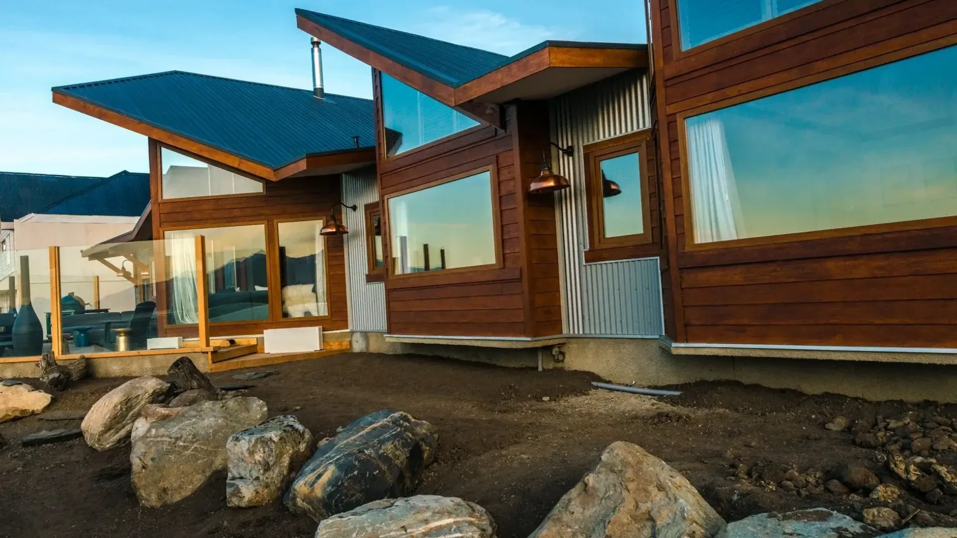 Modern house with large windows and wooden accents, next to a rocky yard under a blue sky.