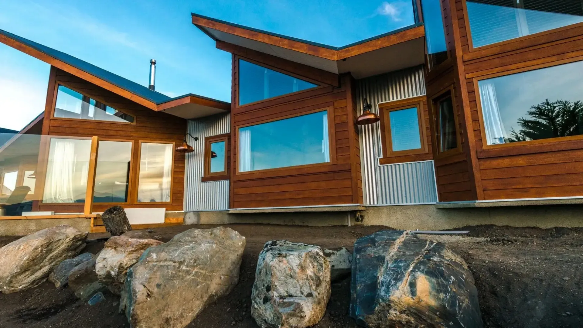 Modern wood and corrugated metal house with large windows, set against a blue sky, rocks in the foreground.