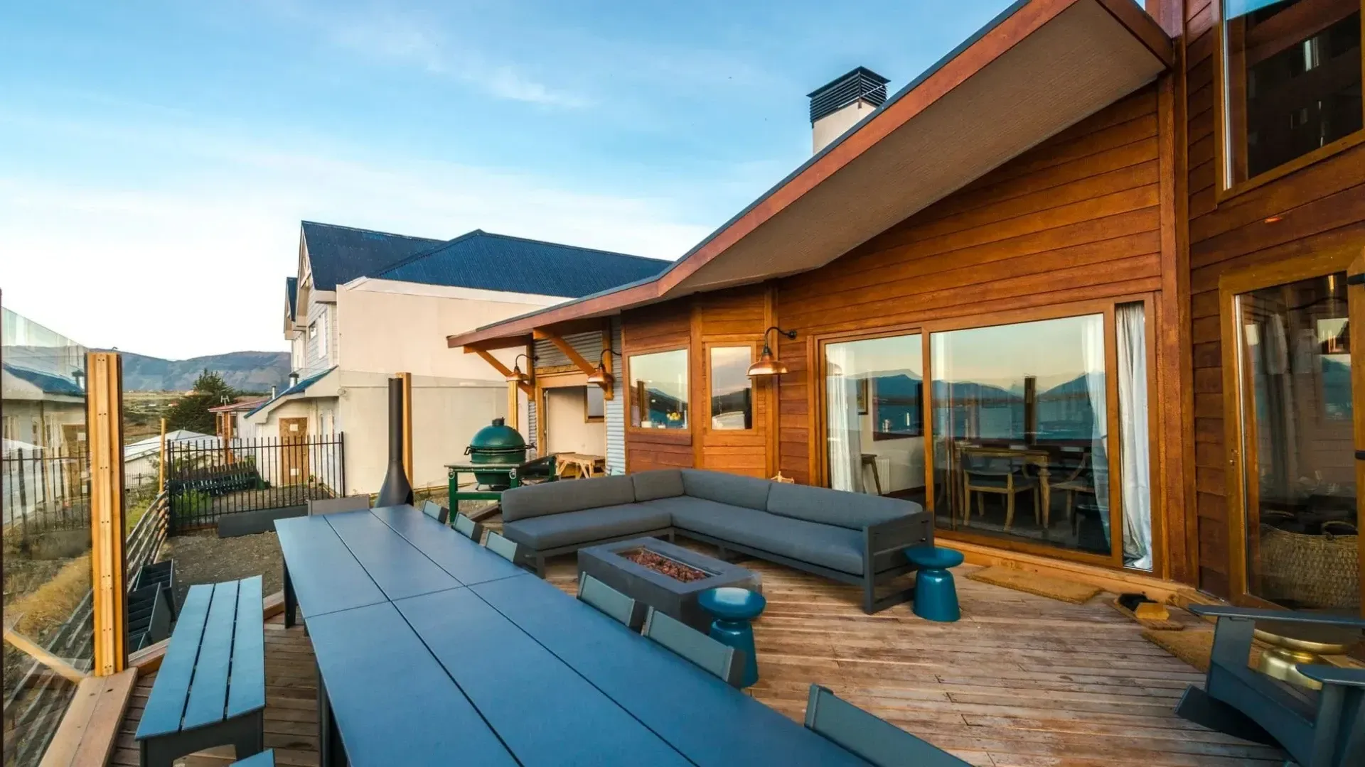 Wooden deck with long blue table and seating, leading to a cabin with a view.