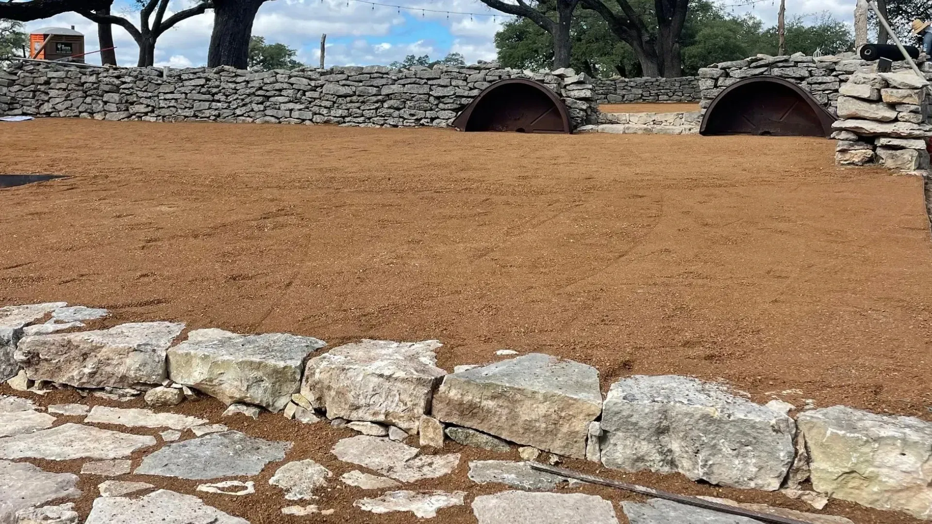 Stone ruins and arches with a freshly tilled area. Large trees in the background under a blue sky.