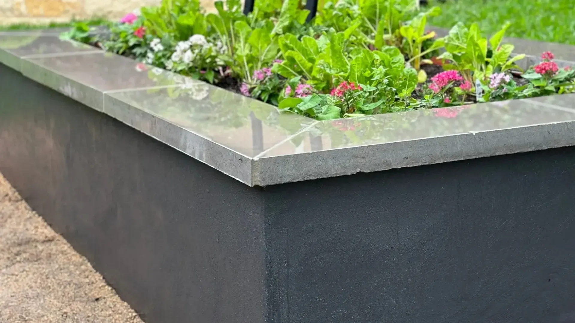 Black planter box with stone coping, filled with green plants and pink flowers, in a garden.