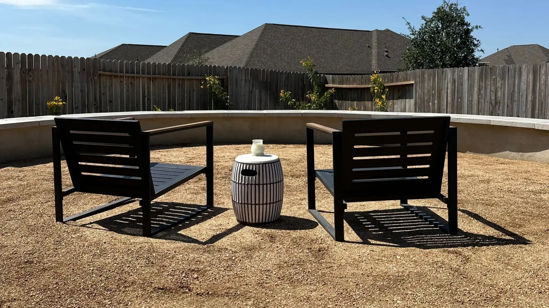 Two black chairs face each other on a gravel patio with a small striped table in between; a wooden fence in the background.