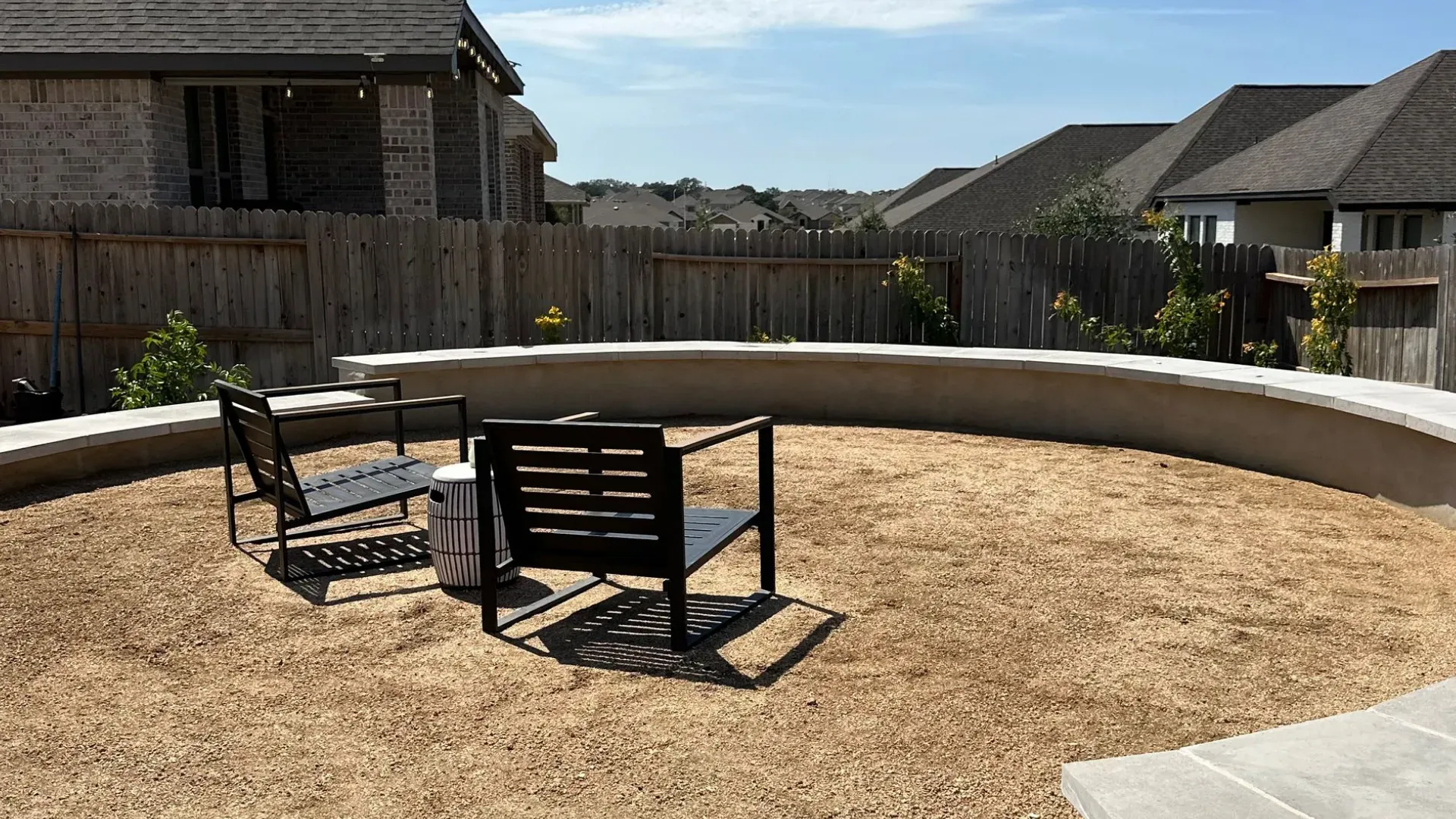 Backyard fire pit area with seating, surrounded by a low wall, under a blue sky.