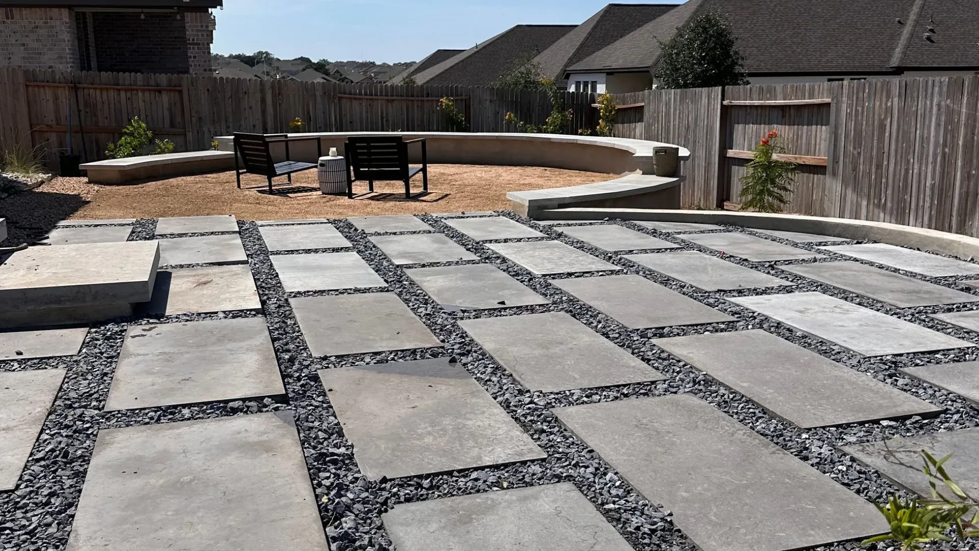 Backyard patio with gray concrete pavers and gravel, under a blue sky.