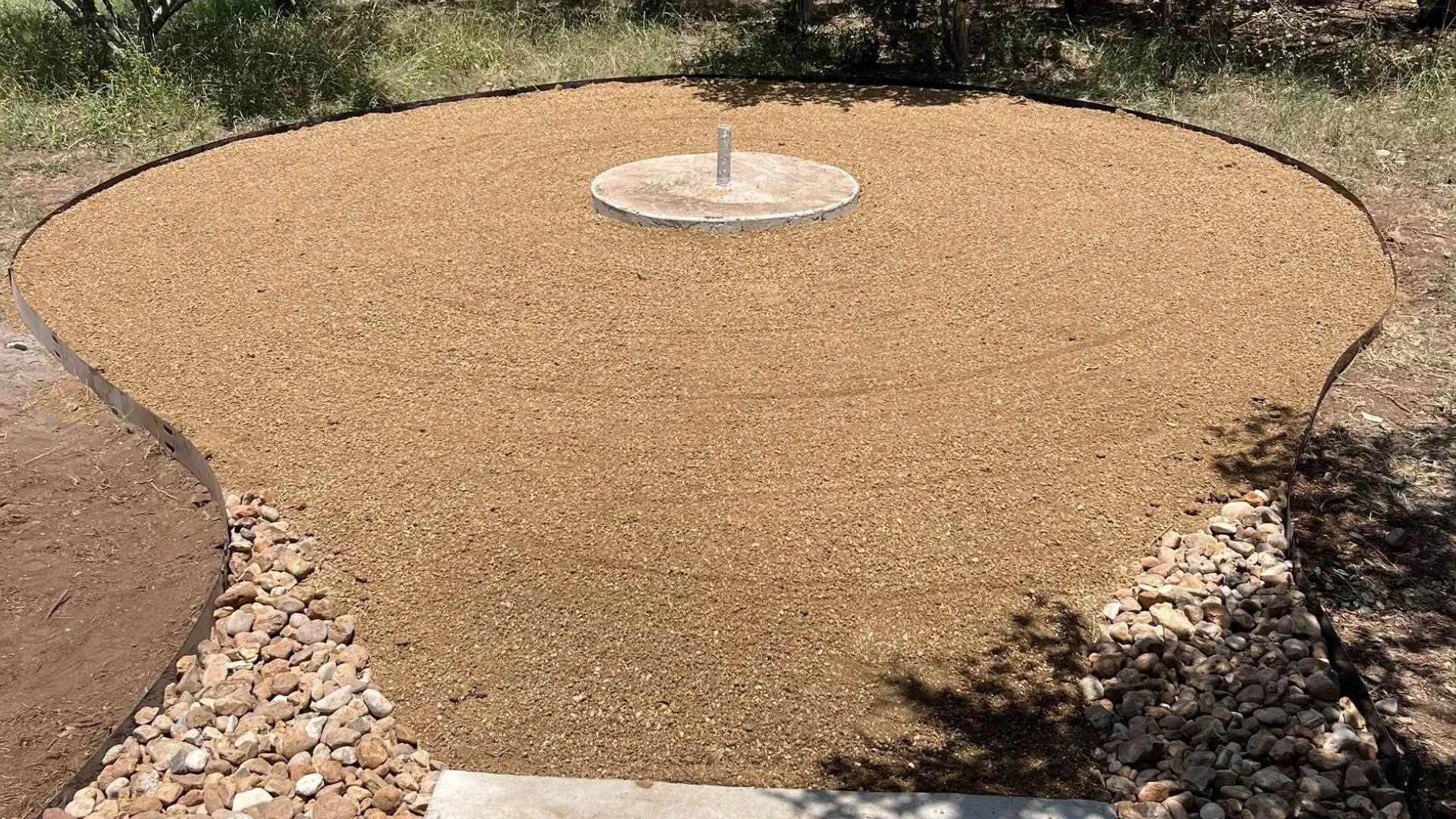 Gravel circle with a fountain, surrounded by trees. Stepping stones lead to the circle. Sunny day.
