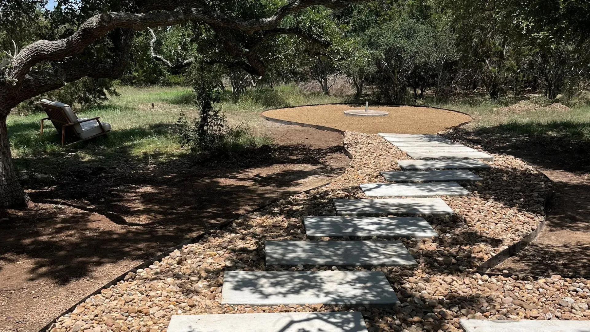 Stone path through a yard, bordered by small brown rocks, under a tree on a sunny day.
