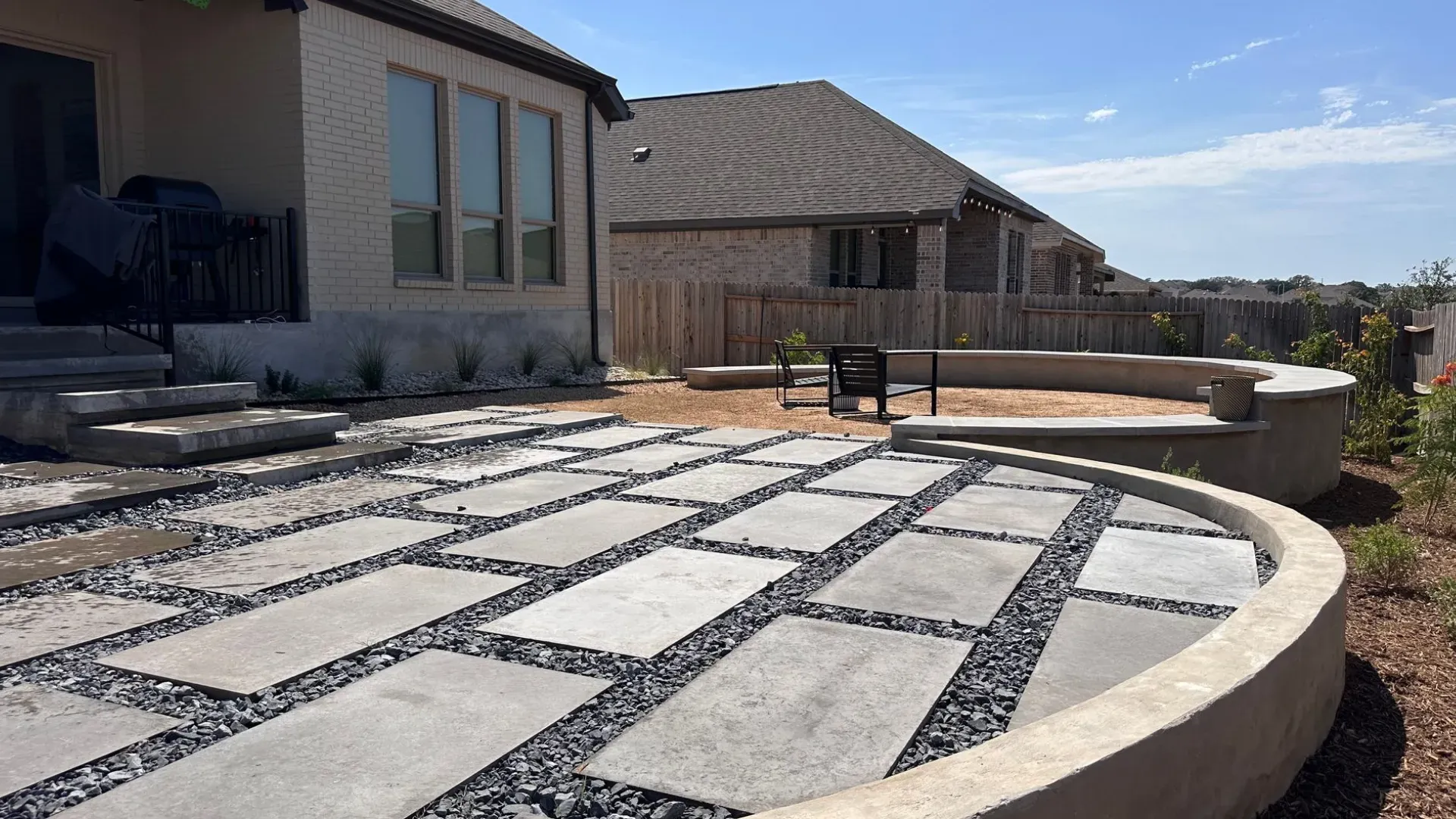 Backyard patio with large gray rectangular pavers and black gravel infill, bordering a curved concrete retaining wall, with a sunny blue sky.