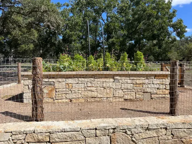 Stone-walled raised garden bed with wire fencing and wooden posts, green plants in the center, trees in the background.