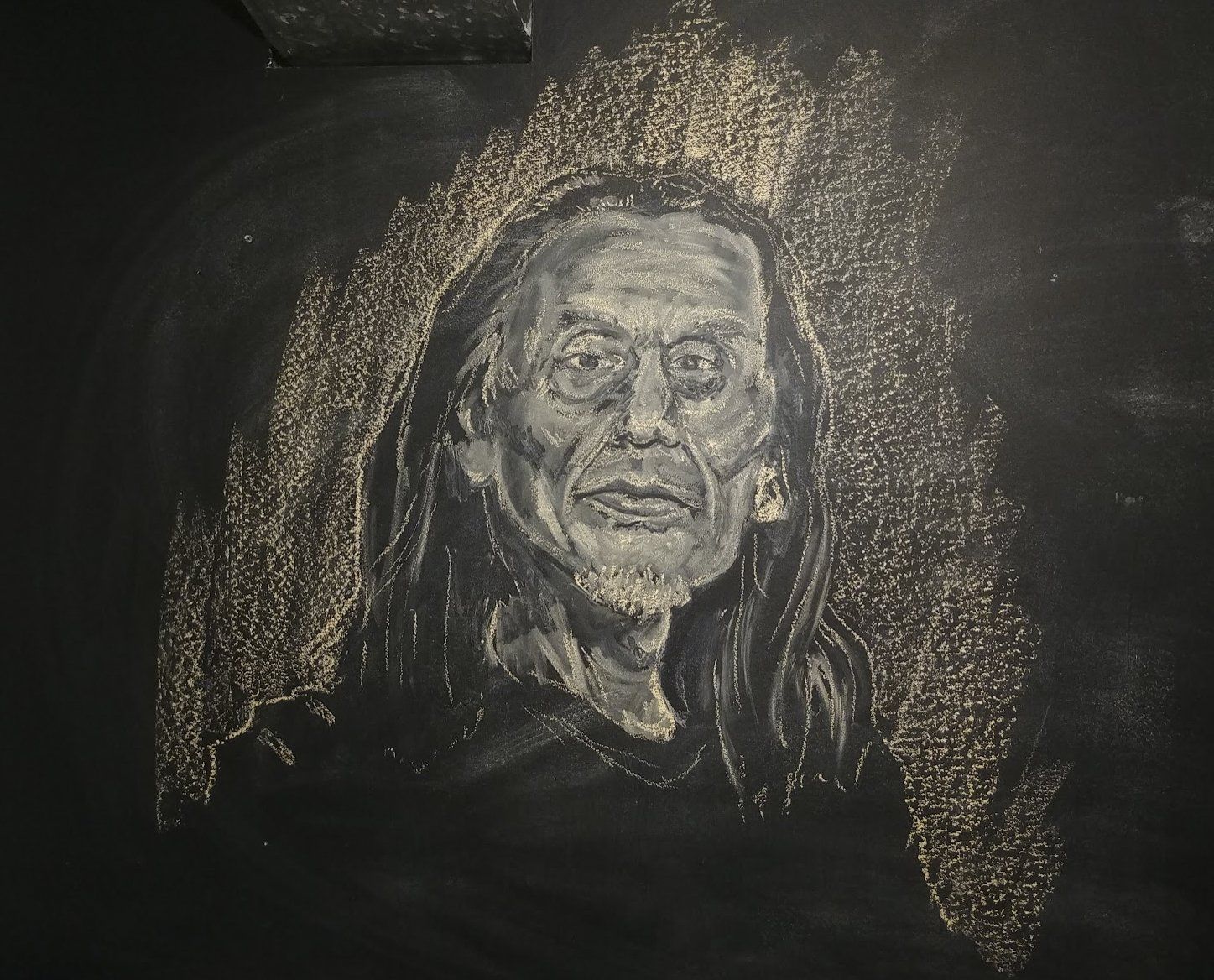 White chalk portrait on black chalkboard; subject with long hair, beard, and focused expression.