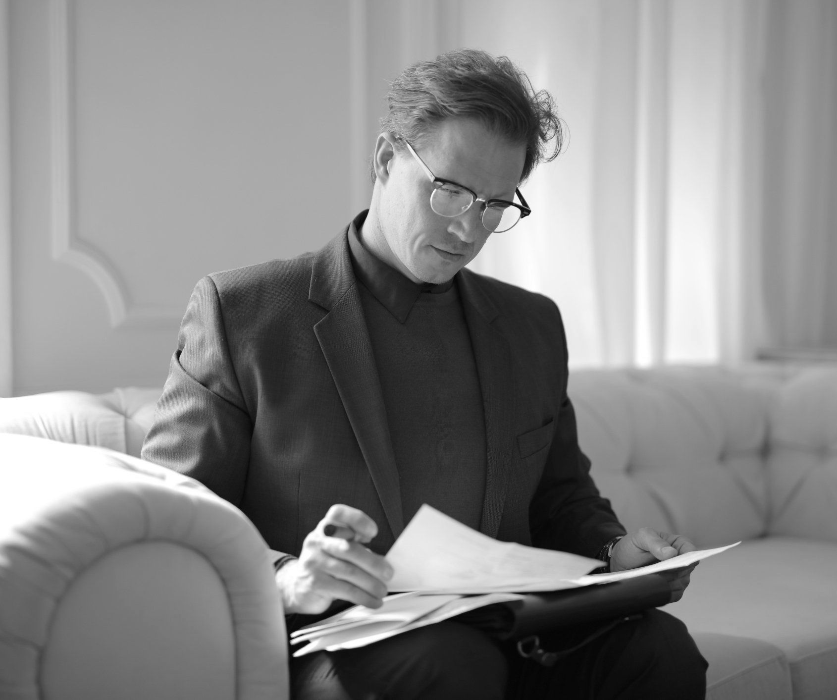 Person in glasses reviews documents on a couch in a bright room.