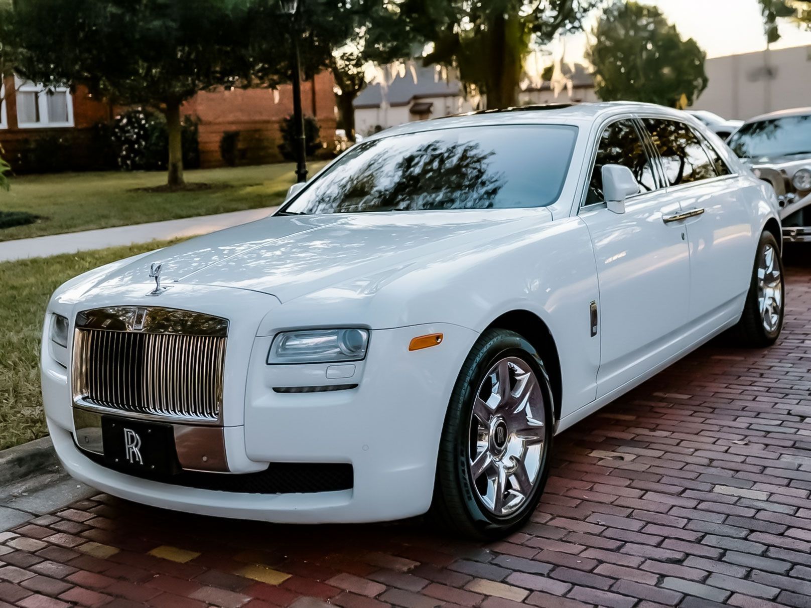 White Rolls-Royce car parked on a brick street near a sidewalk and trees; sunny daylight.
