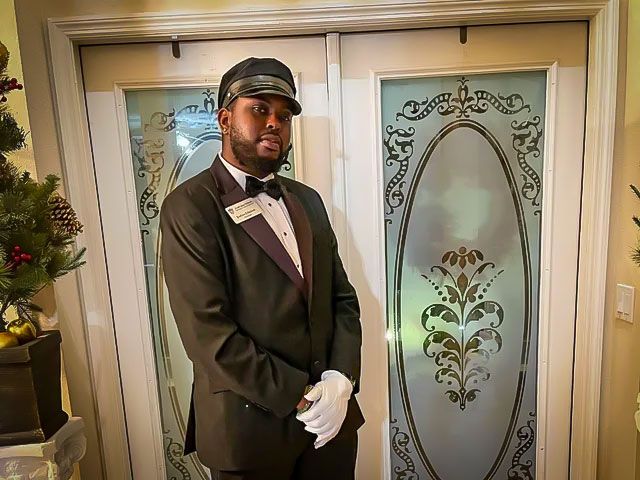 Doorman in formal attire stands by glass doors with ornate designs. He wears a hat and gloves.
