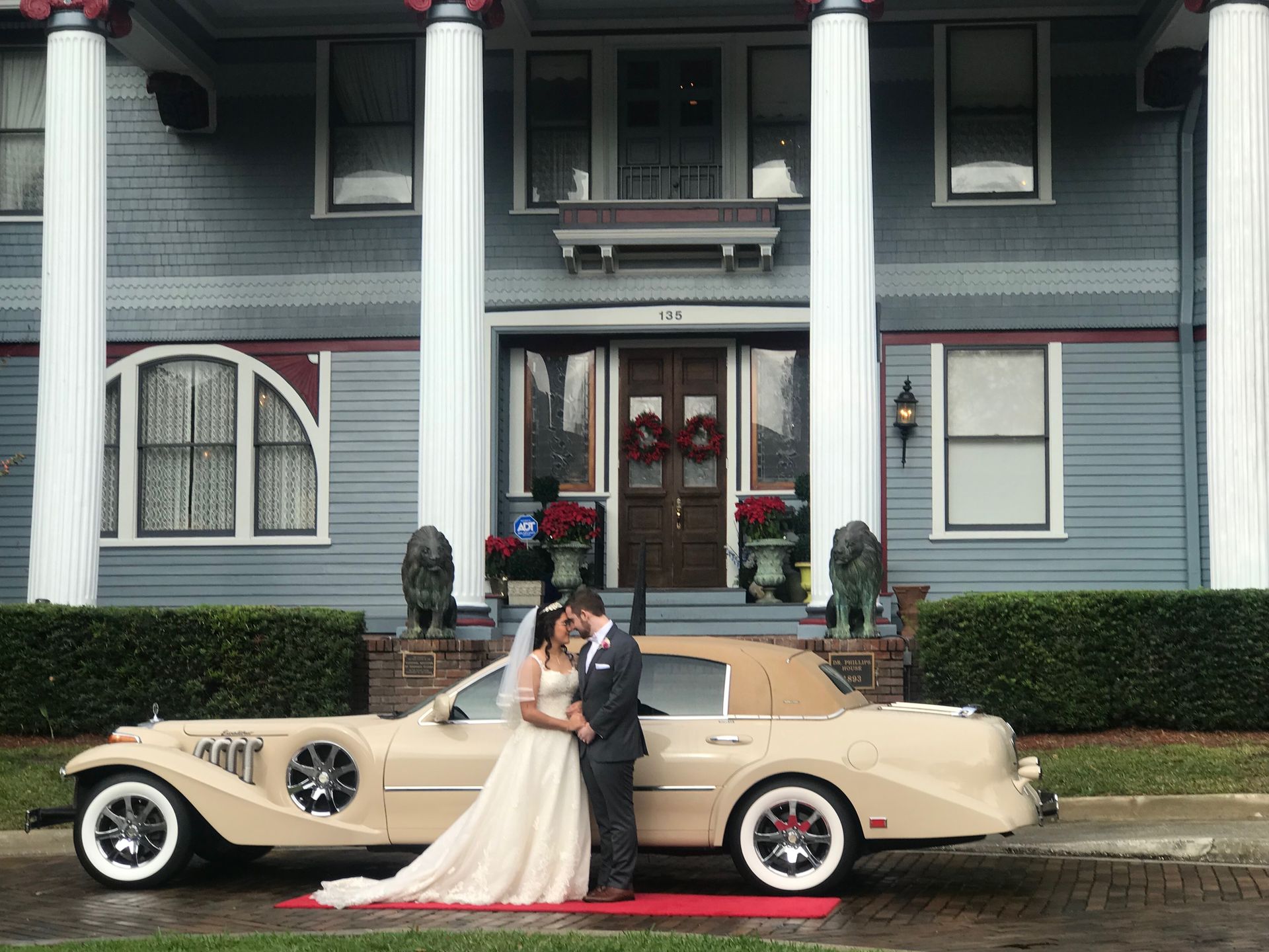 Exotic Limo Orlando Couple embraces beside a beige convertible in front of a blue mansion with white columns; a red carpet leads up to the door.