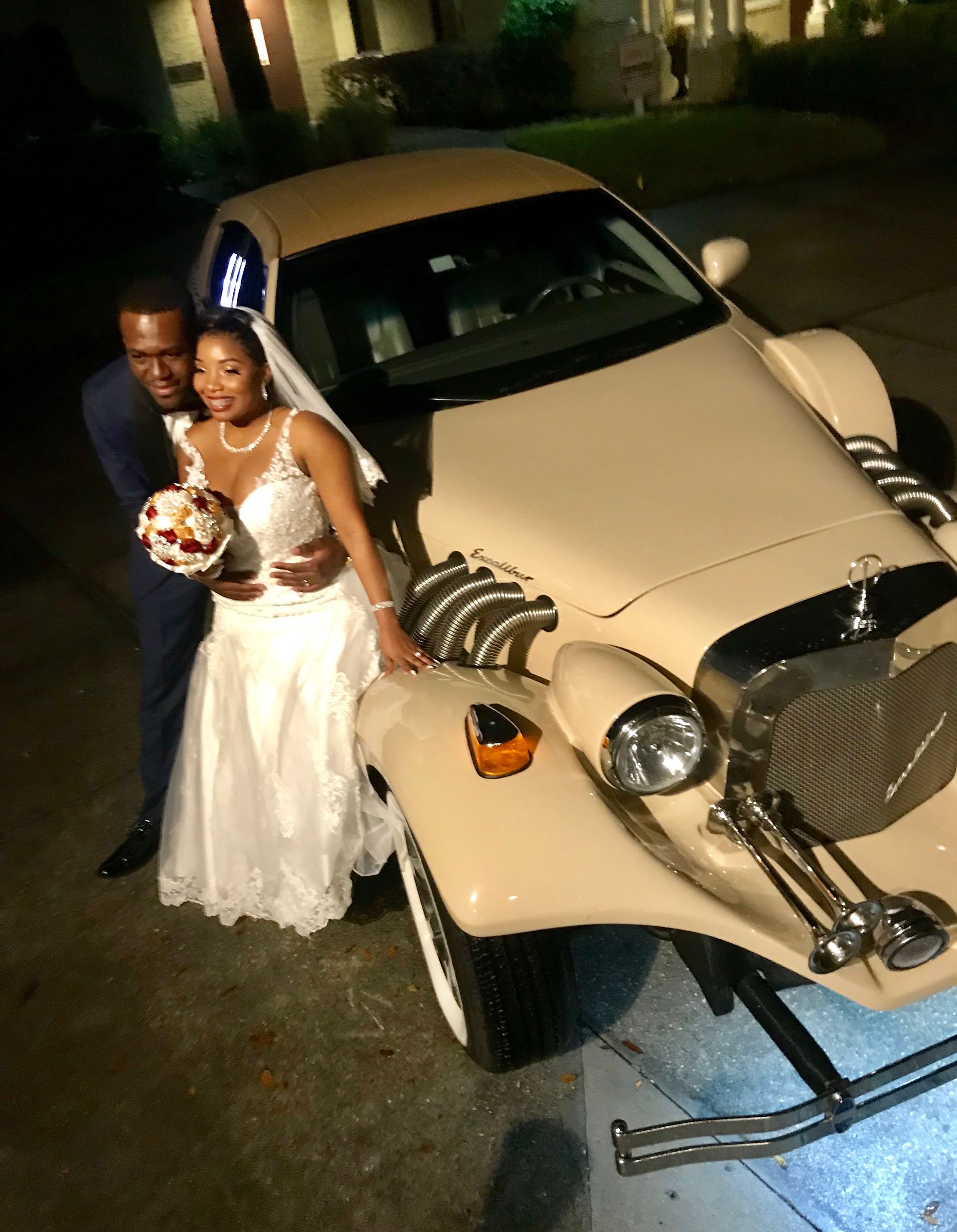 Exotic Limo Orlando Wedding couple poses by vintage cream-colored car at night. Bride wears white gown, groom wears navy suit.