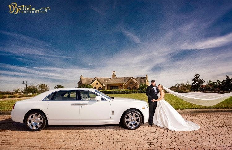Exotic Limo Orlando Wedding couple poses near white Rolls-Royce with flowing veil; blue sky background, brick road.