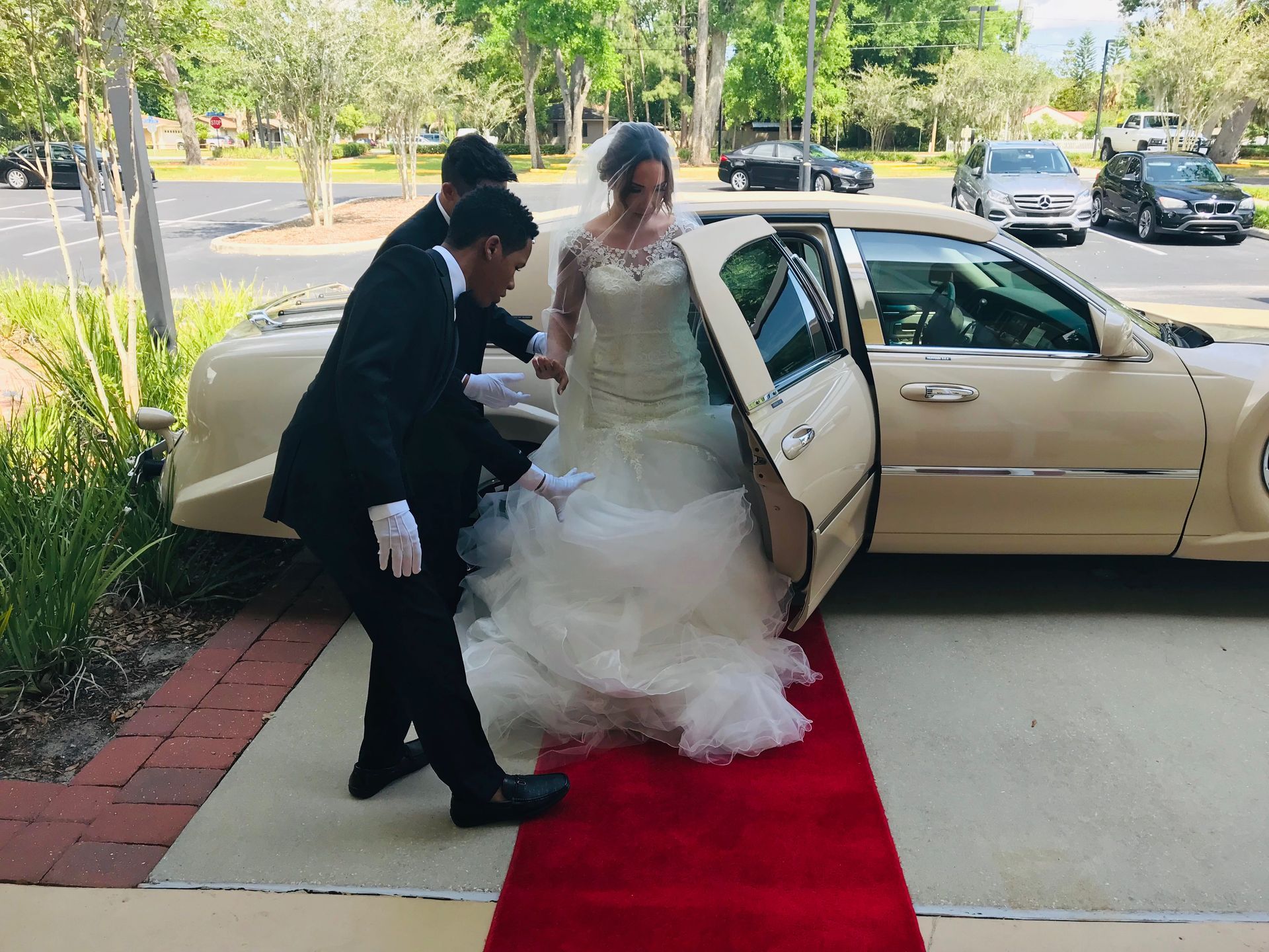 Exotic Limo Orlando Bride exiting a cream-colored limousine, assisted by two men, onto a red carpet. Sunny outdoor setting.