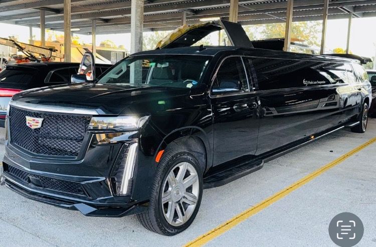 Man in tuxedo and gloves leans against a black car, looking away. City background. Exotic Limo Orlando