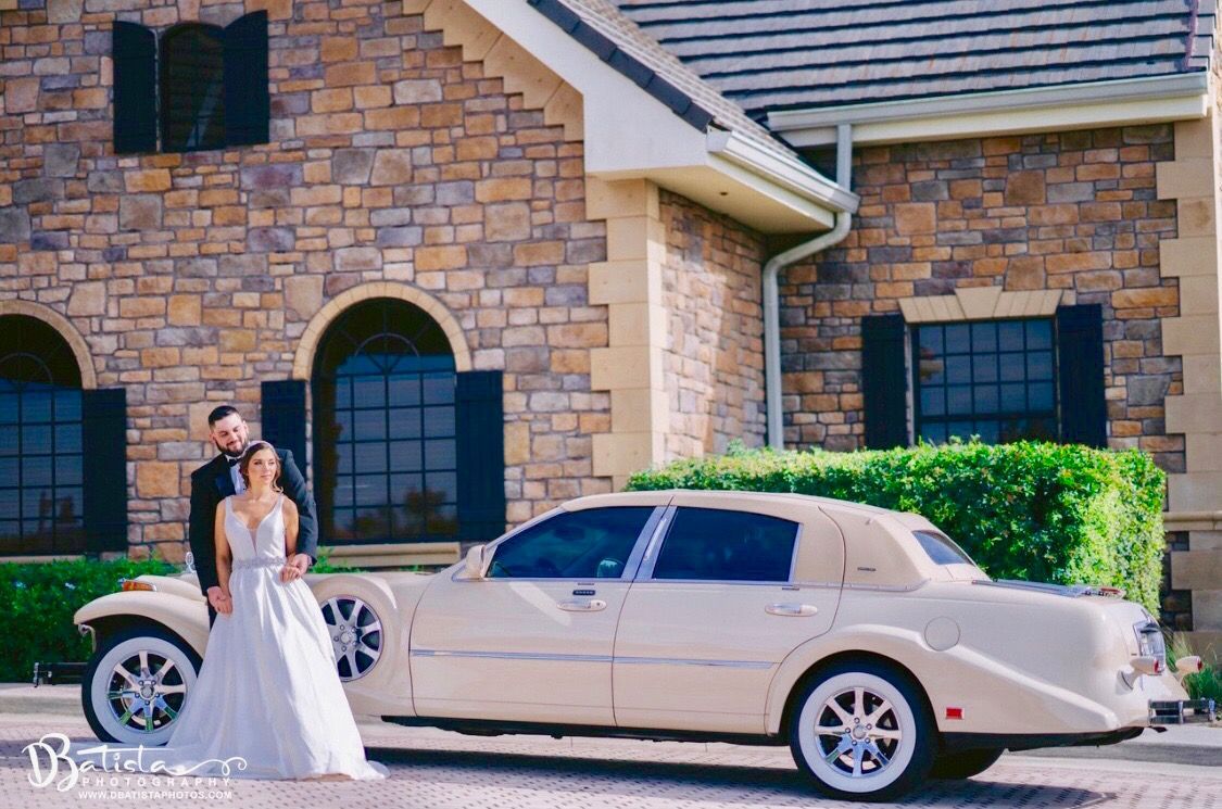 Bride and groom pose by a cream limousine in front of a stone building with black shutters.