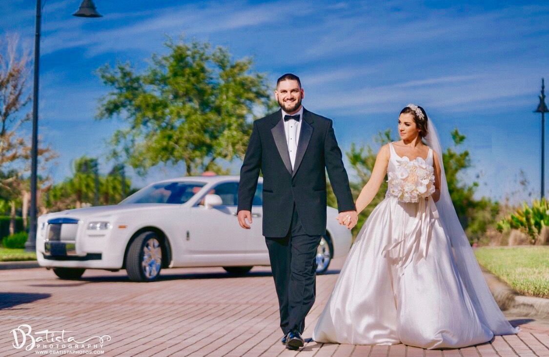Bride and groom holding hands, walking towards the camera. White Rolls Royce parked nearby, sunny day.Exotic Limo Orlando
