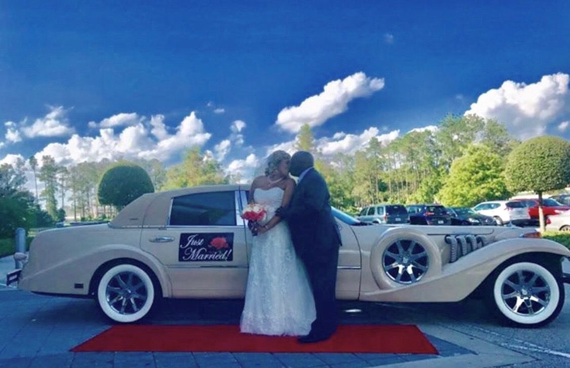 Exotic Limo Orlando Newlyweds kissing beside a vintage car on a red carpet. Sunny day with a blue sky and trees.