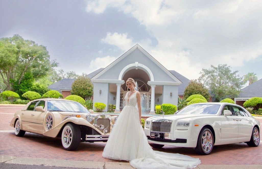 Bride in a wedding dress stands between a vintage car and a modern Rolls Royce, in front of a building.Exotic Limo Orlando