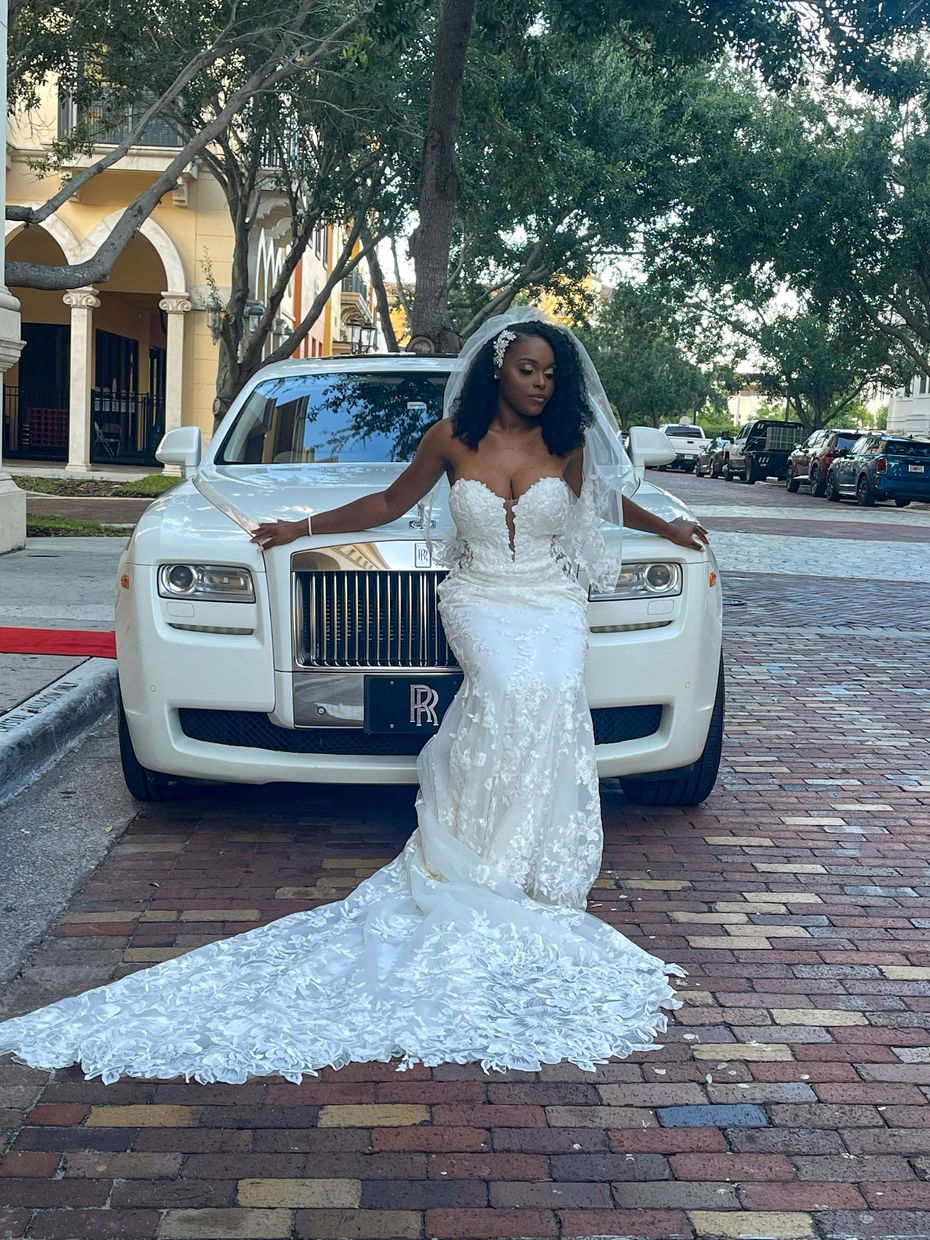Bride in a white lace wedding gown with a train, poses in front of a white Rolls Royce.