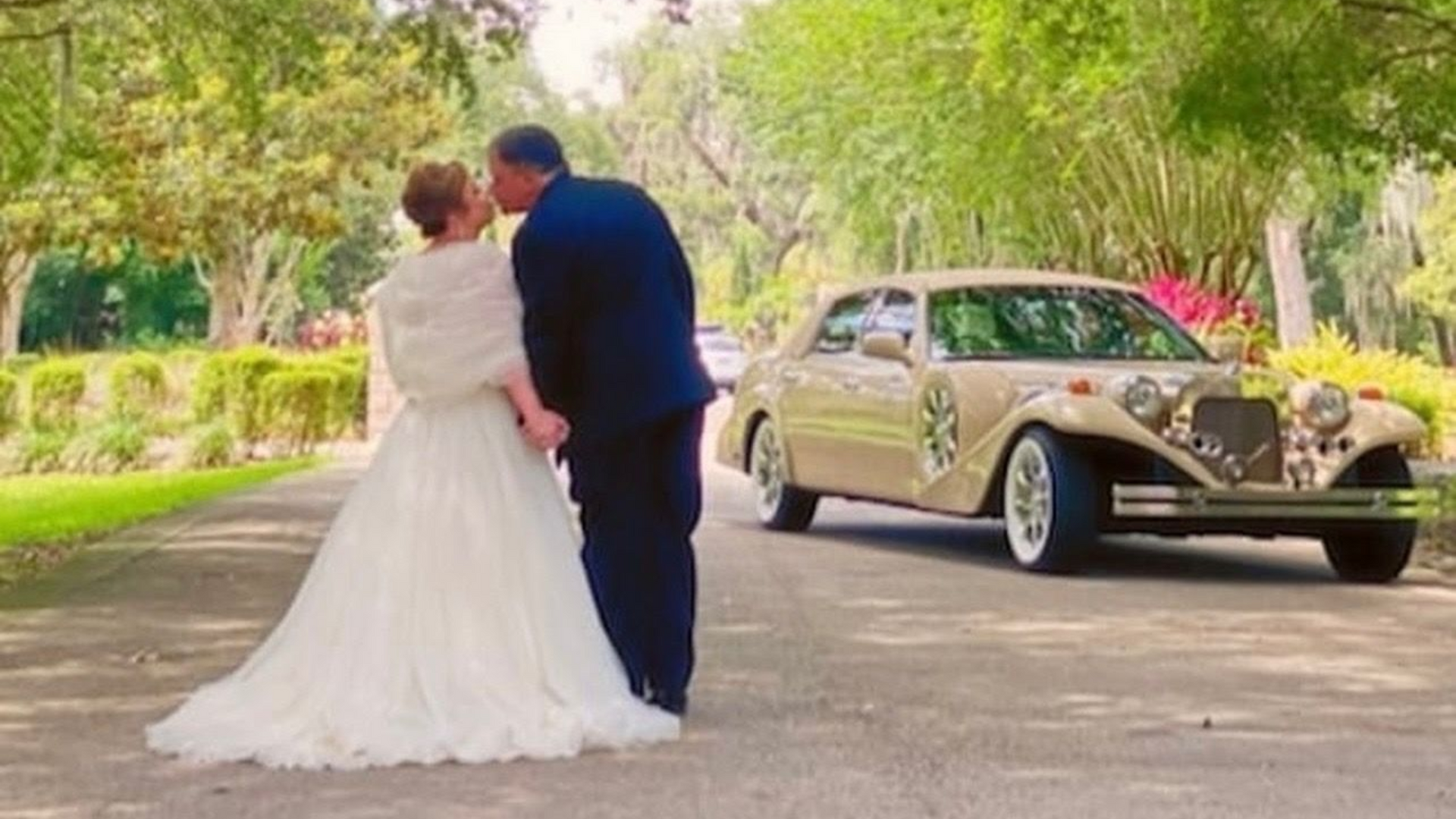 Newlyweds embrace near a vintage beige car on a tree-lined road. Excalibur Godfather Exotic Limo Orlando