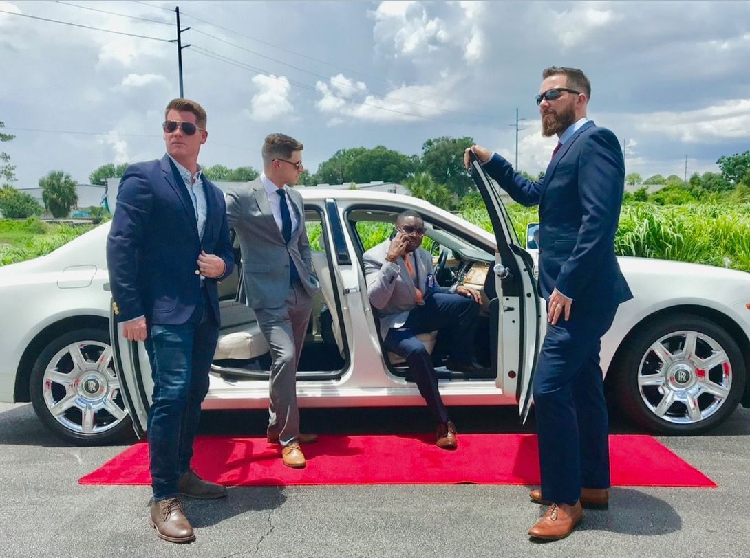 Four men in suits pose near a white car on a red carpet outdoors.