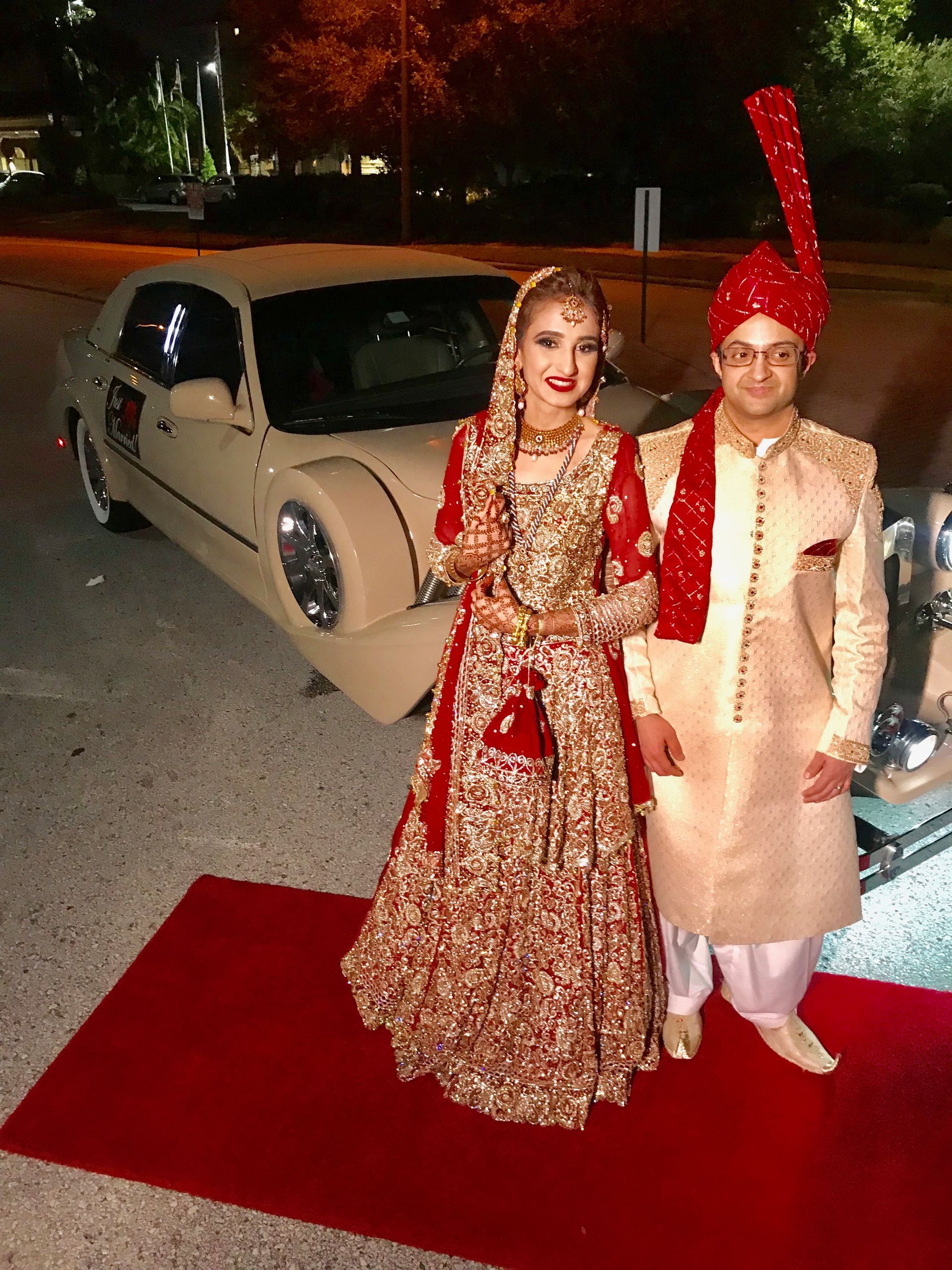 Exotic Limo Orlando Bride and groom in traditional Indian wedding attire stand on a red carpet near a vintage car.