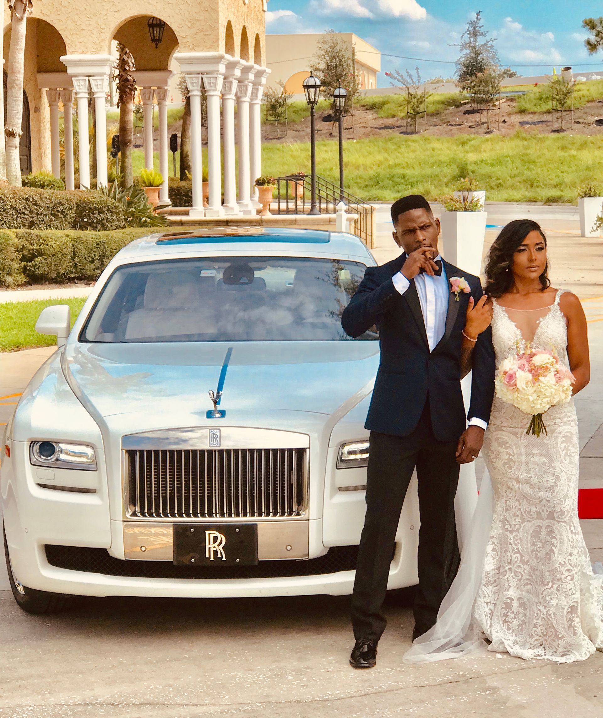 Newlyweds posing next to a white Rolls Royce. The groom wears a suit, and the bride holds a bouquet, outdoor setting.Exotic Limo Orlando